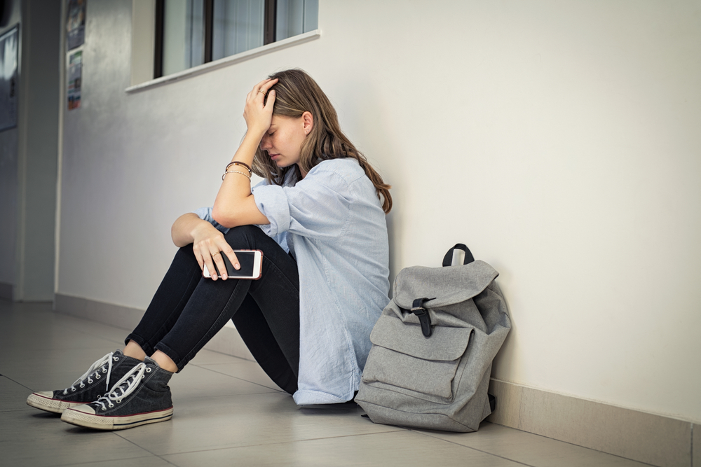 Upset and depressed girl holding smartphone sitting on college campus floor holding head. University sad student suffering from depression sitting on floor at high school. Lonely bullied teen.